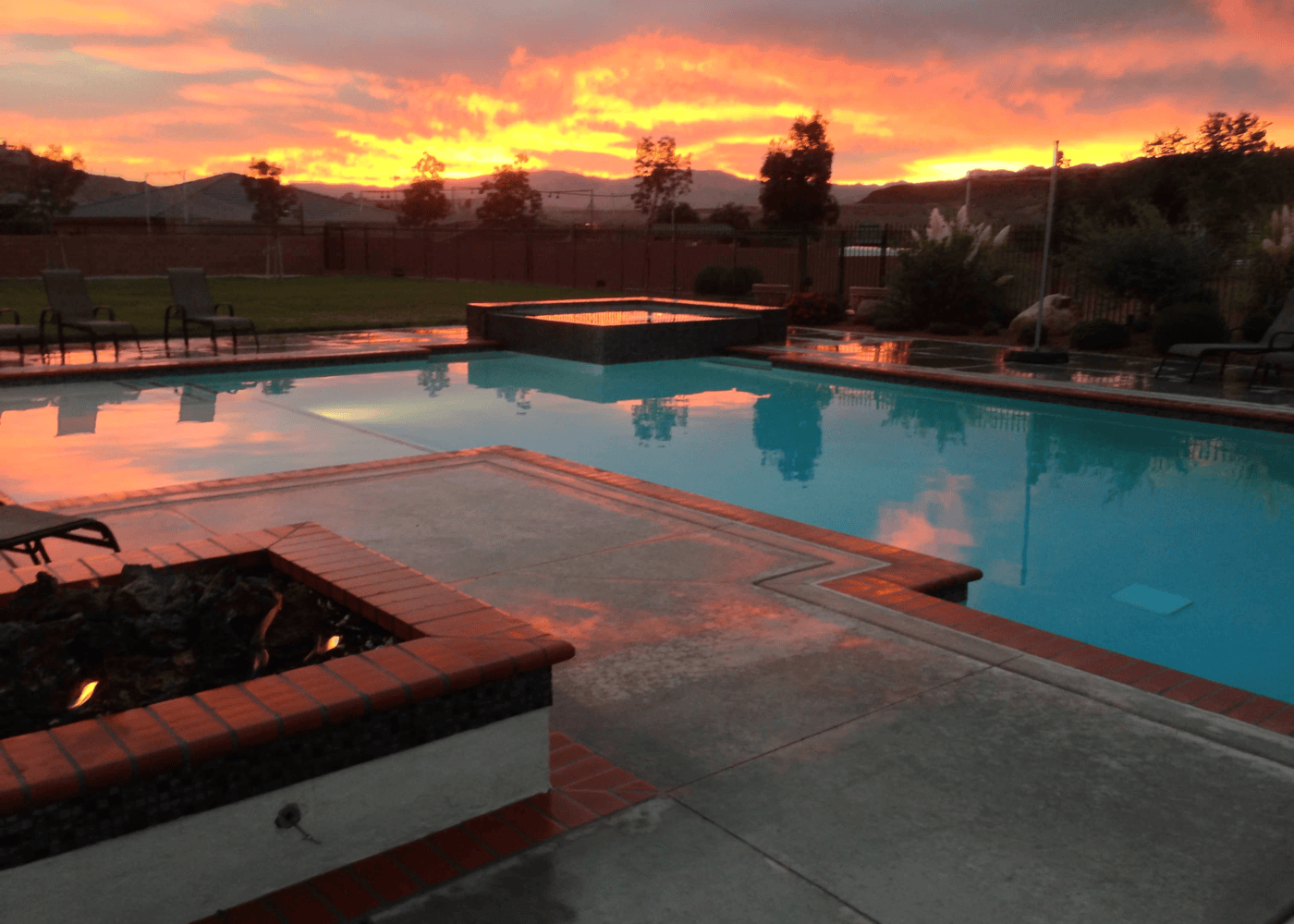 An aesthetic view of an in-ground swimming pool in the evening, showing the sunset, with a firepit in the foreground and the rest of the backyard in the background. The pool is illuminated, casting a gentle glow on the water's surface with the pool's lights.