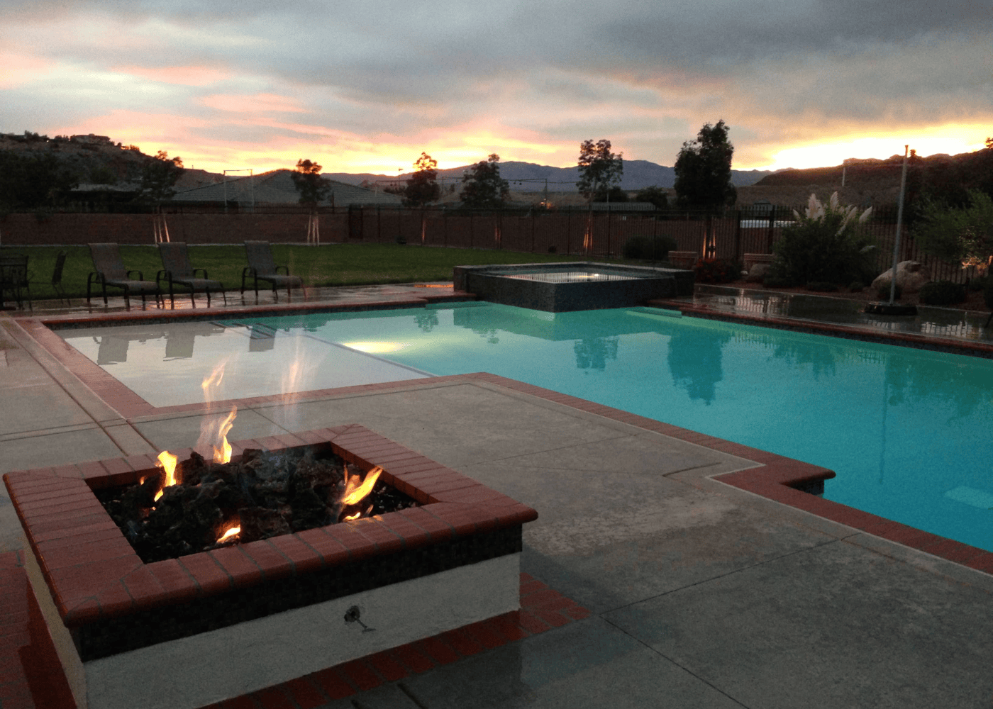 An aesthetic view of an in-ground swimming pool in the evening, with a firepit in the foreground and the rest of the backyard in the background. The pool is illuminated, casting a gentle glow on the water's surface with the pool's lights.
