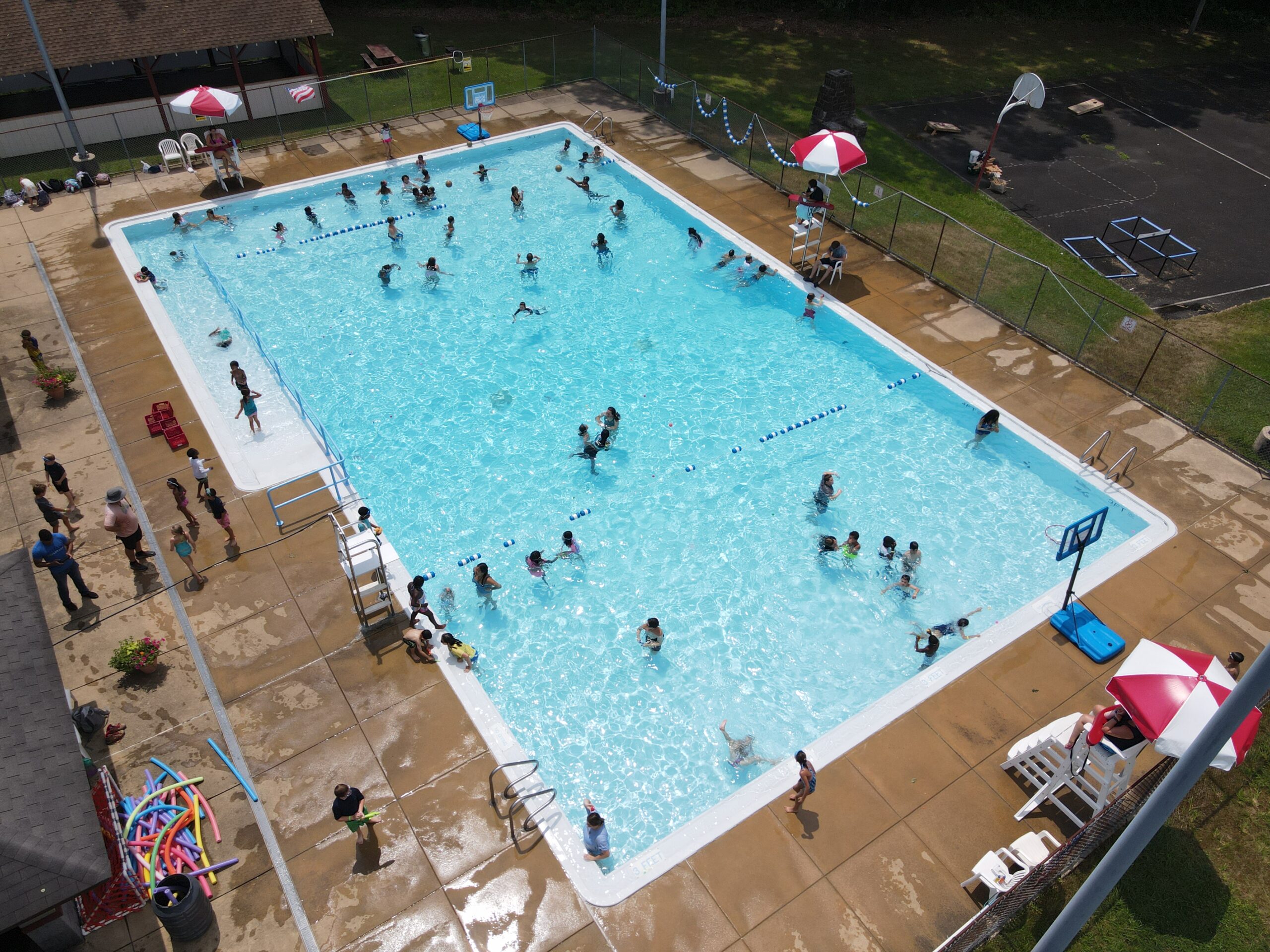 A view of a commercial in-ground swimming pool coated with ecoFINISH, showcasing the entire pool and its surrounding area. The surrounding area includes people enjoying the pool, some swimming and others lounging poolside.