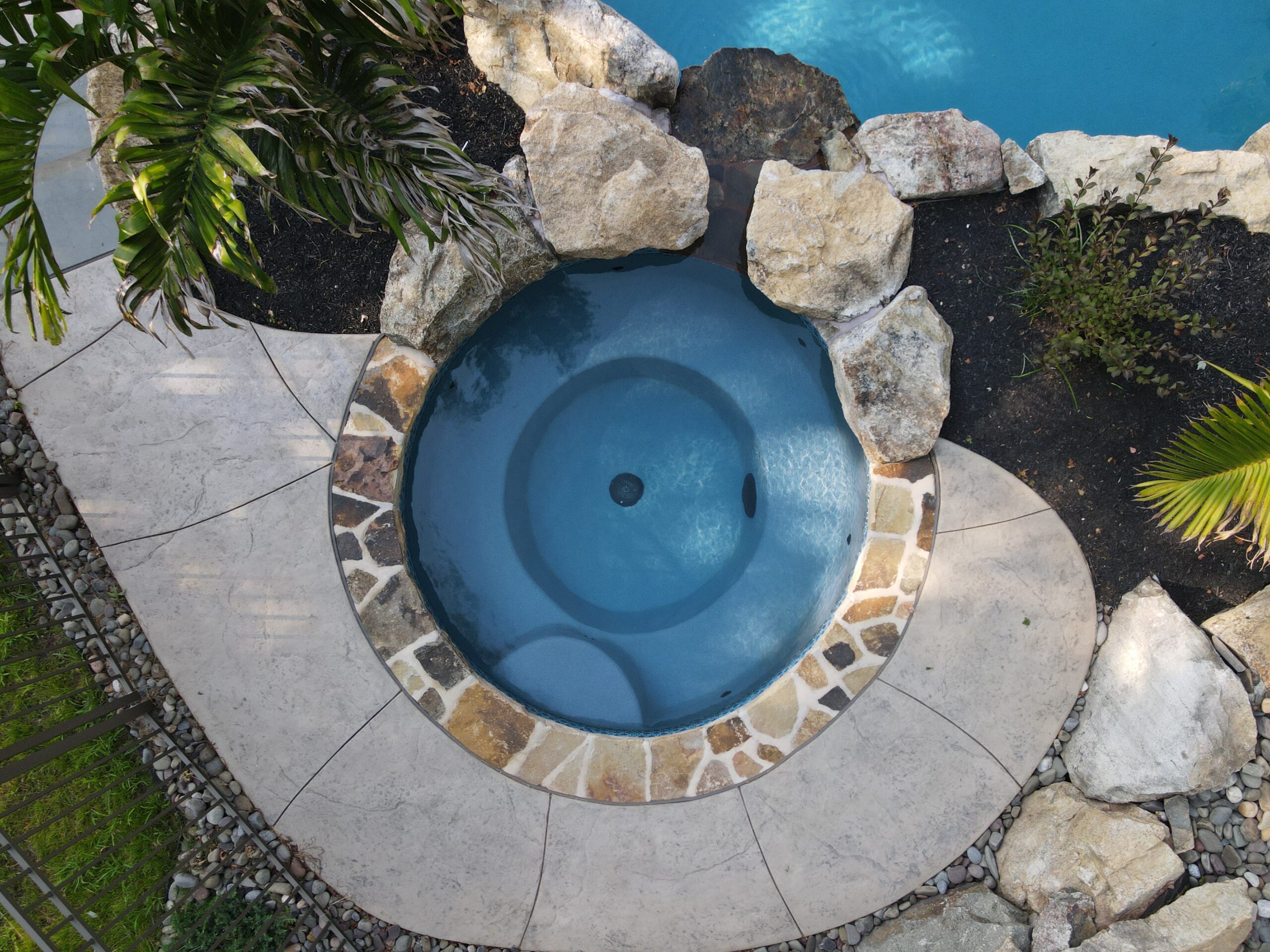 A bird's-eye view of a spa connected to a swimming pool, with rocks surrounding the spa area. The spa features clear water, while the rocks enhance the natural aesthetic of the poolside environment.