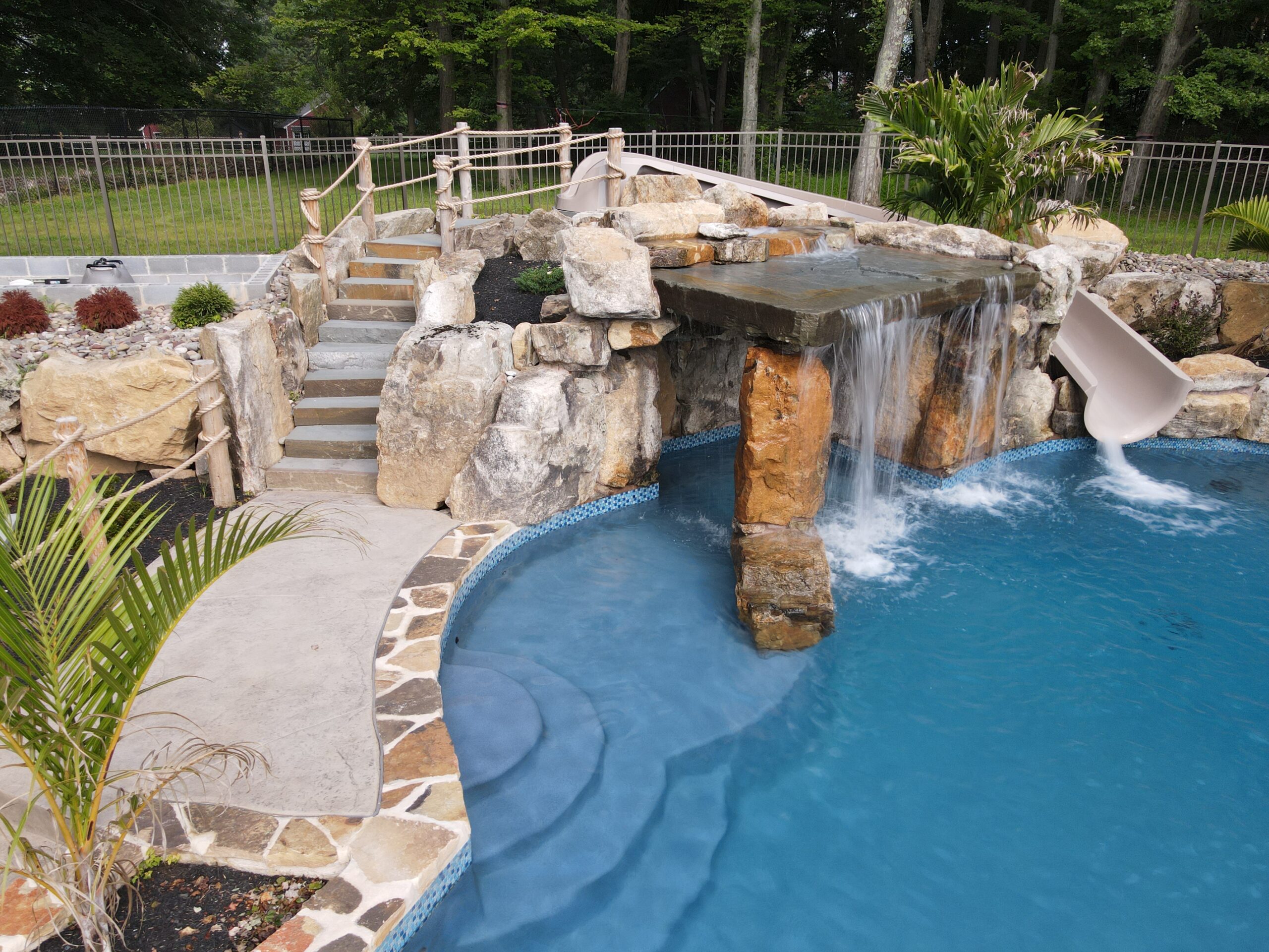 An ecoFINISH coated pool with a waterfall grotto cascading into clear water, and steps leading up to a slide at the far end of the pool.