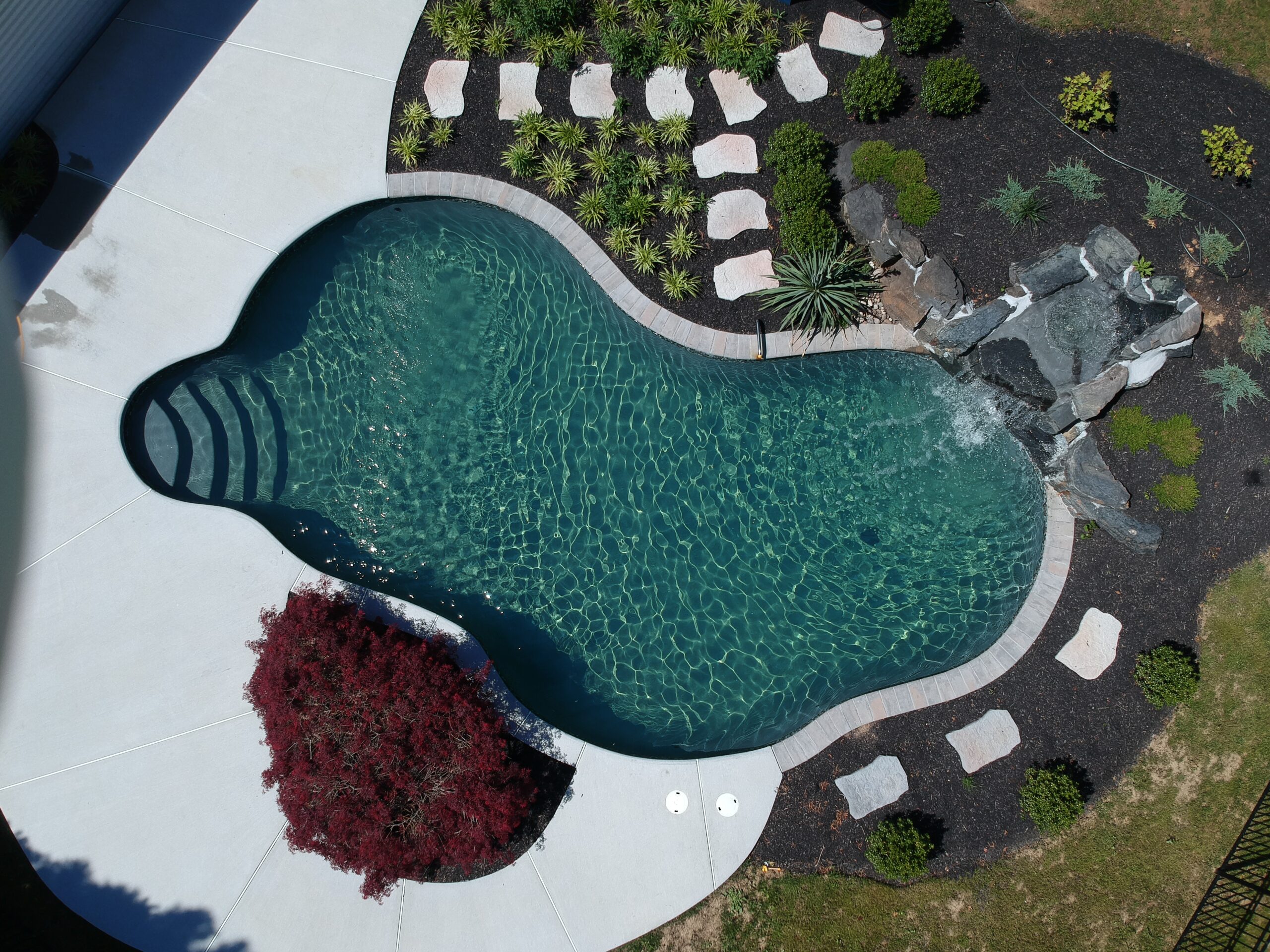 A bird's-eye view of an ecoFINISH-coated pool, showcasing the pool's surface and the surrounding landscape. At the end of the pool, a waterfall cascades into the water, adding a natural and scenic element to the setting.