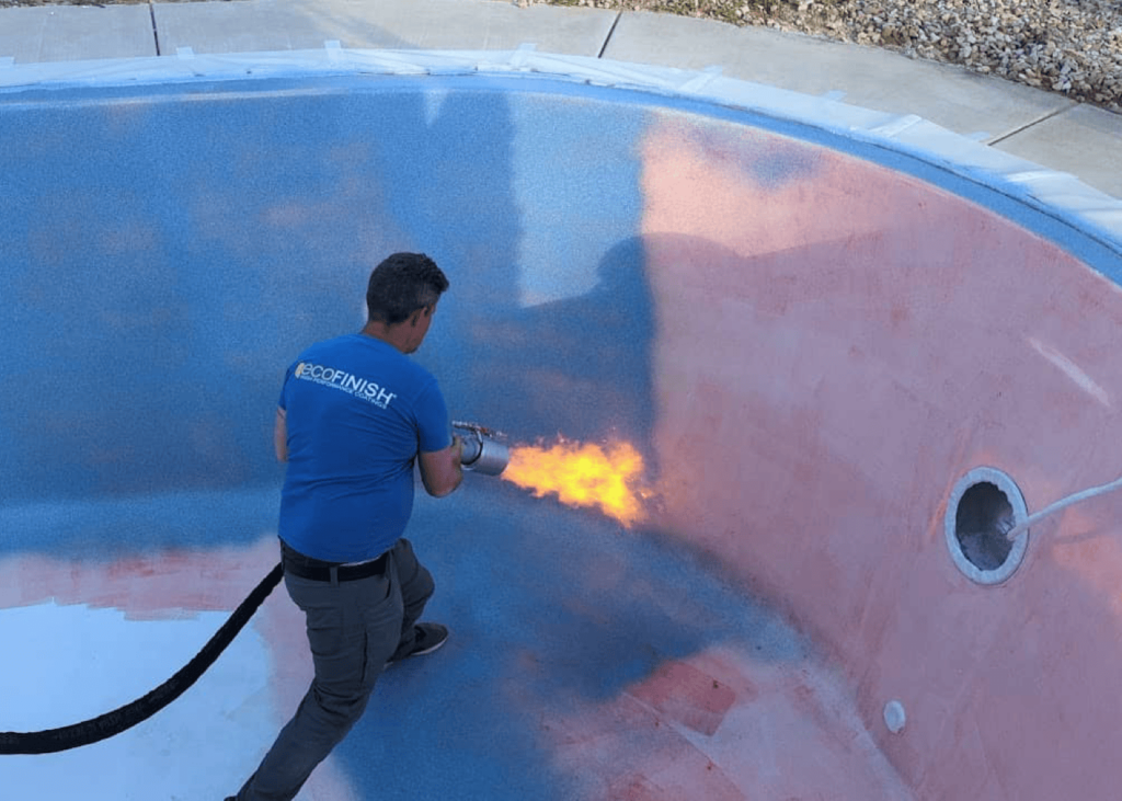 A construction worker wearing a blue shirt with the "ecoFINISH" logo is inside an empty, oval-shaped swimming pool. He is using a large heat torch to apply a new finish. The pool is partially a light blue speckled color and partially a pinkish-red primer.