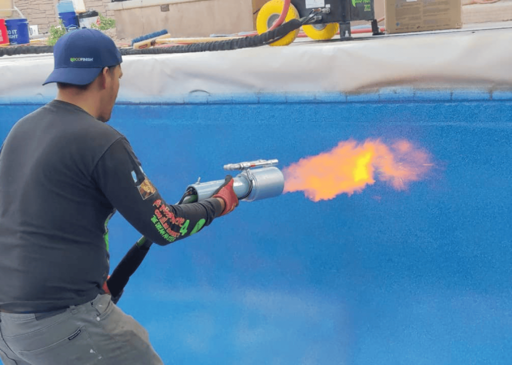 A worker, seen from behind, is using a large heat torch to apply a new finish to the interior of a swimming pool. The tool emits a large orange flame directed at the bright blue, textured pool surface.