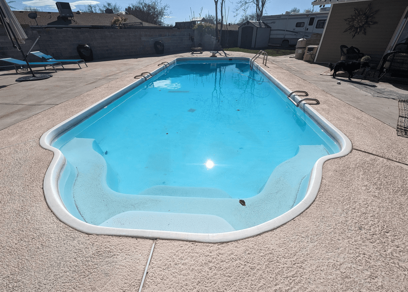 An in-ground pool filled with clear water, reflecting sunlight. The pool's serene surface contrasts with the deck's textured paving stones.