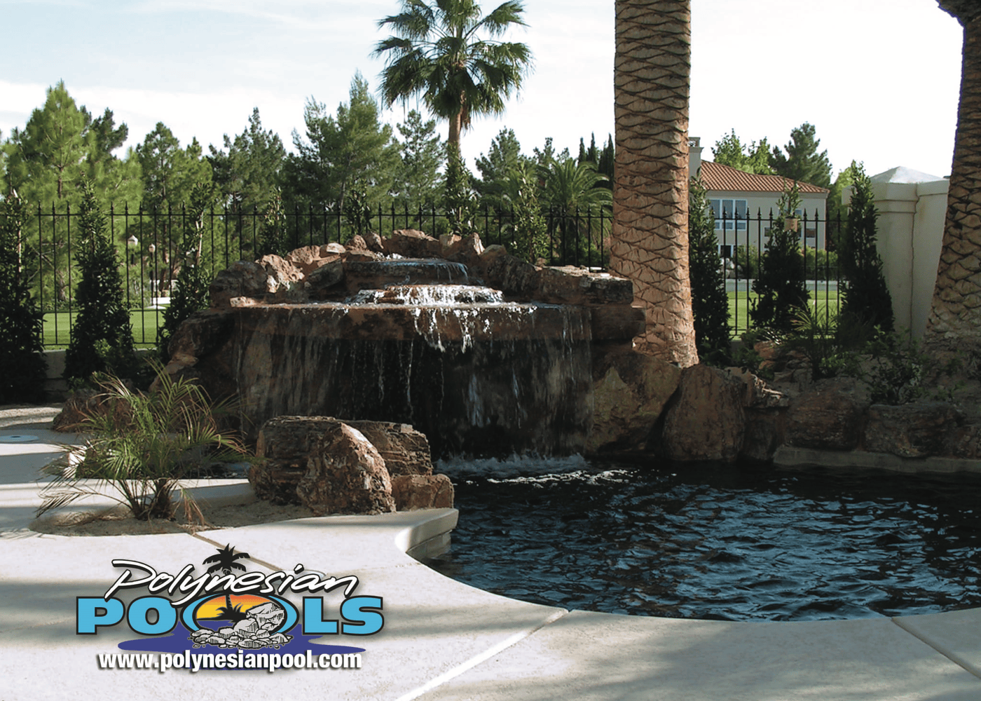 A view of an in-ground swimming pool showcasing a waterfall/grotto feature at one end. Surrounding landscaping and the pool's dark blue water enhance the relaxing ambiance.