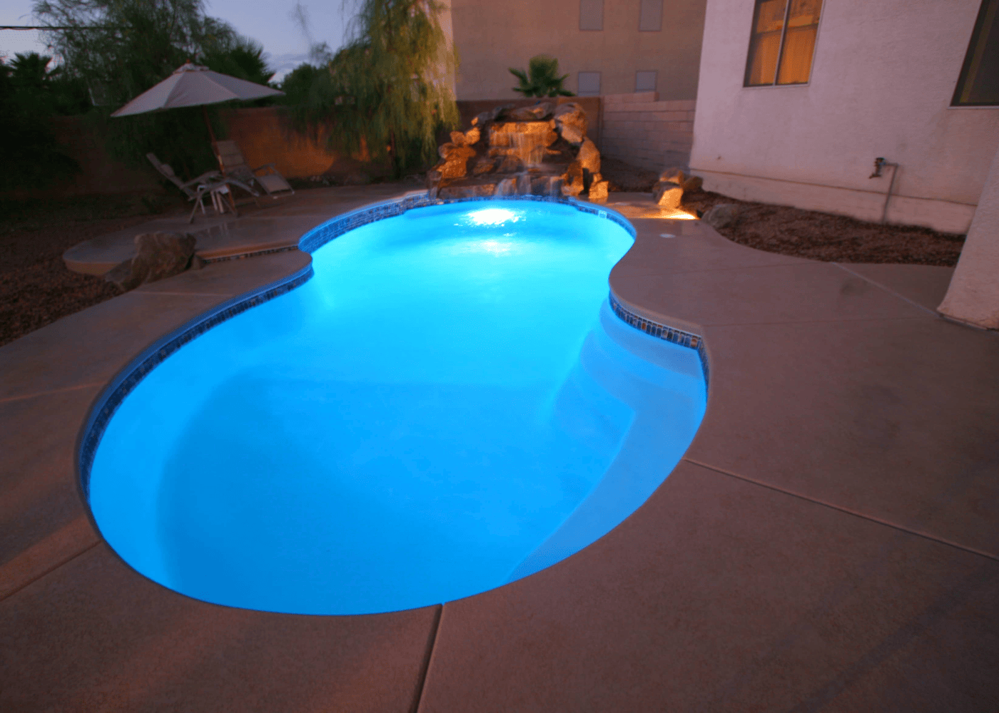 A filled in-ground swimming pool illuminated in the evening with the pool lights on. The clear blue water reflects the ambient lighting, creating a tranquil and inviting atmosphere.