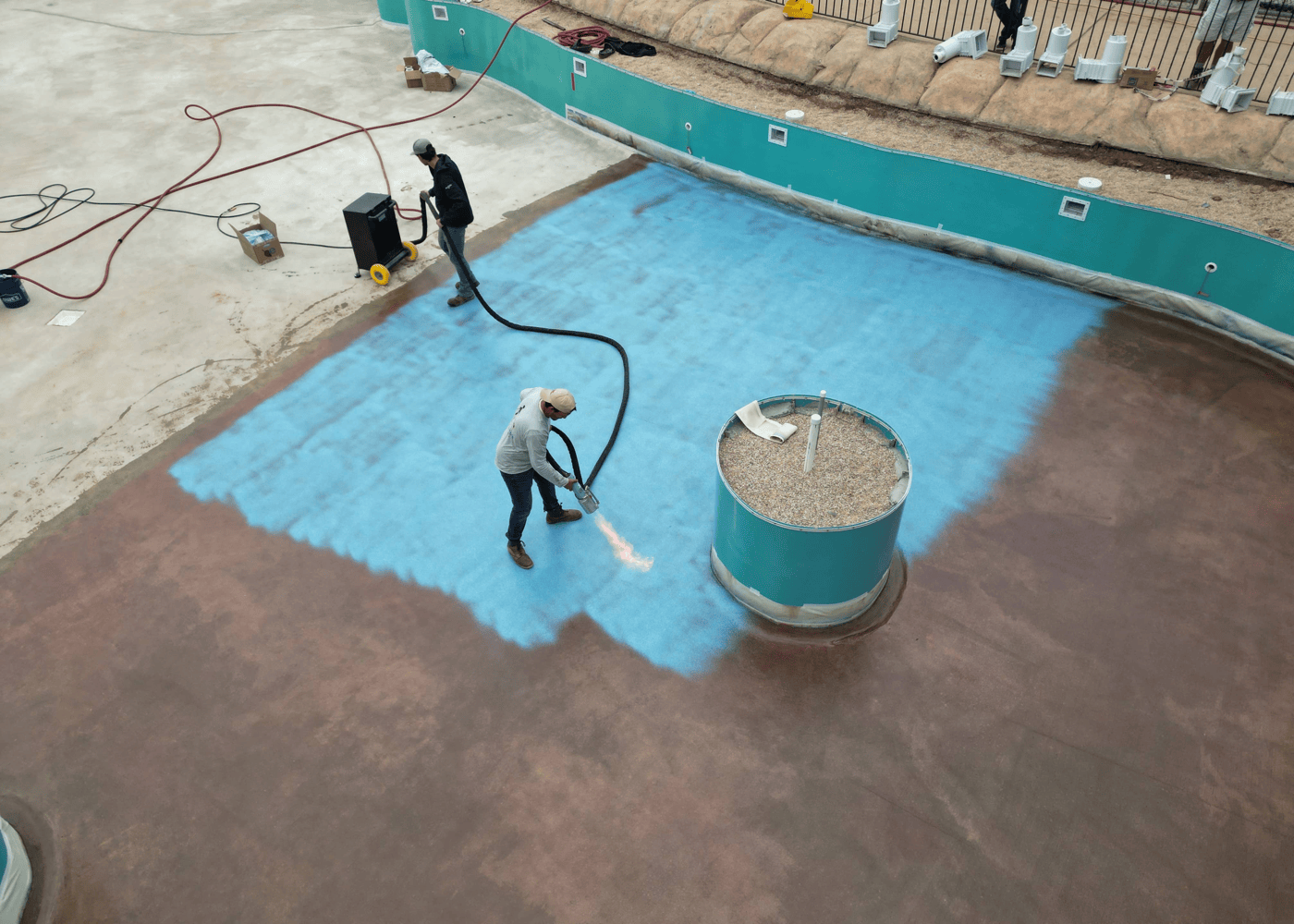 A view of an empty in-ground swimming pool in the process of being coated with ecoFINISH. Workers are applying the ecoFINISH coating material to the pool's interior surface.