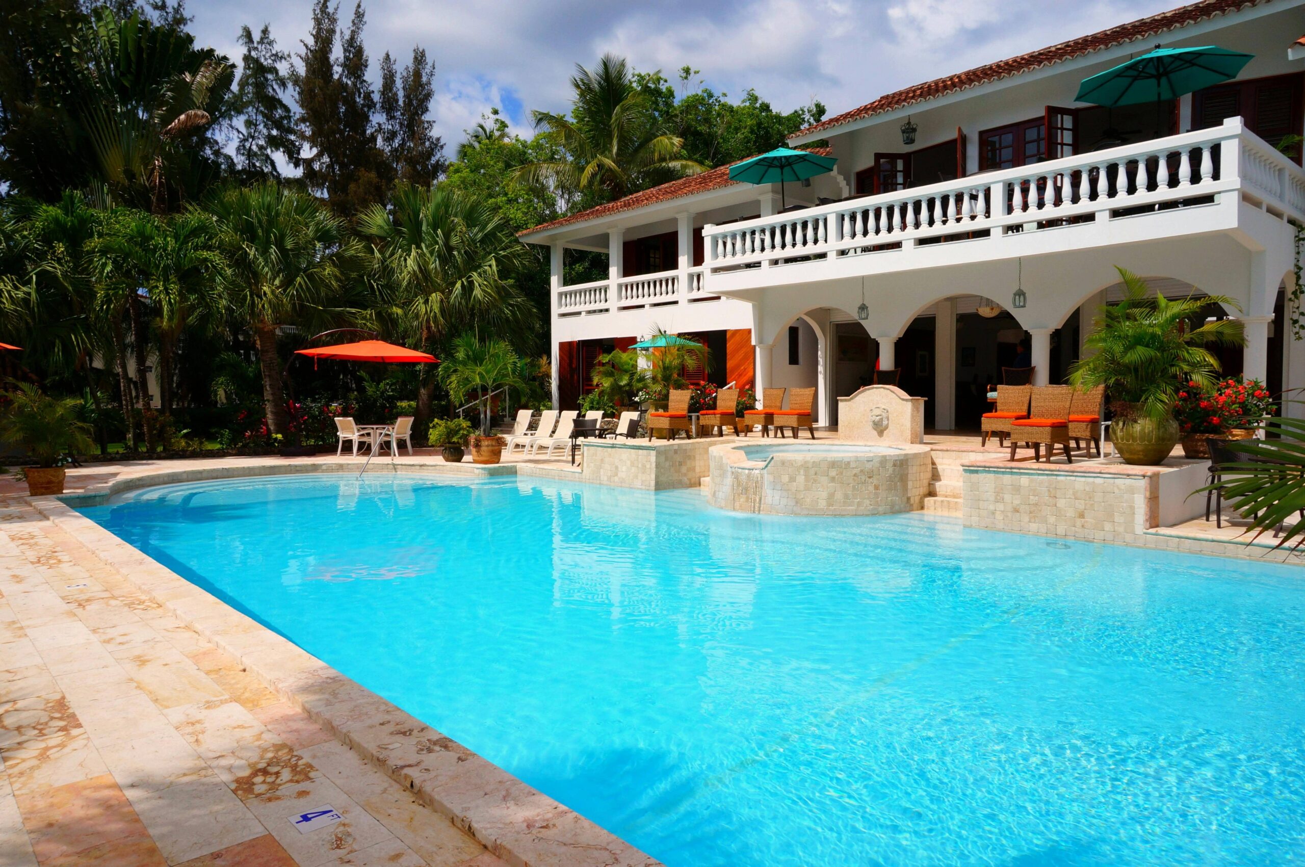 A bright, tropical resort-style swimming pool and multi-story building. The pool has a vibrant blue interior and a light-colored tiled deck with lounge chairs and umbrellas. There is a small, raised spa with water spilling into the main pool.