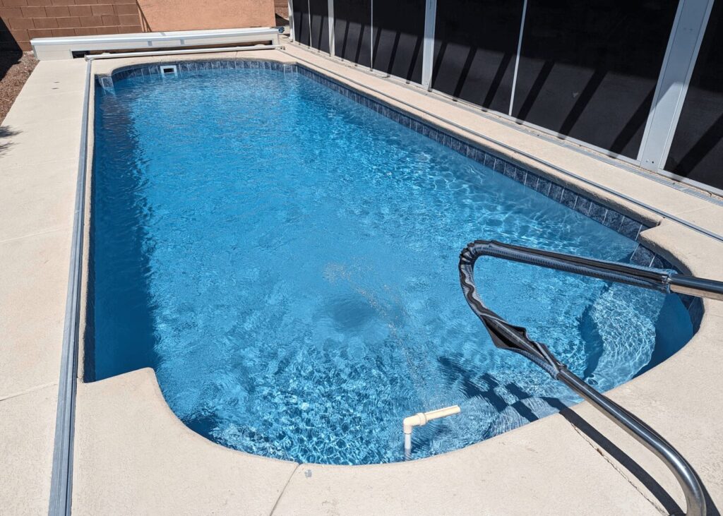 A high-angle shot of a light blue swimming pool with curved ends and decorative tiling along the waterline. A metal handrail with a black protective cover sits in the foreground, and a white automatic pool cover housing is visible at the far end.