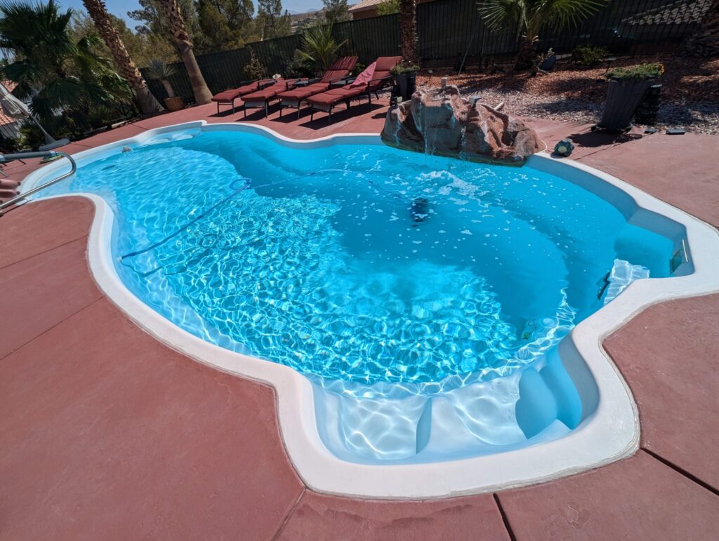 A wide-angle view of a curvy, bright blue backyard swimming pool surrounded by a reddish-brown patio. A faux-rock waterfall feature sits on the edge of the pool, and several red lounge chairs are lined up in the background under palm trees.