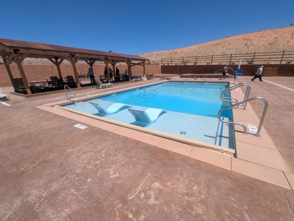 An outdoor swimming pool with clear blue water under a bright, sunny sky. The pool features a shallow "sun shelf" area with two light blue lounge chairs partially submerged in the water. To the left, a large wooden pavilion with a corrugated metal roof provides shade over several lounge chairs and a group of people.